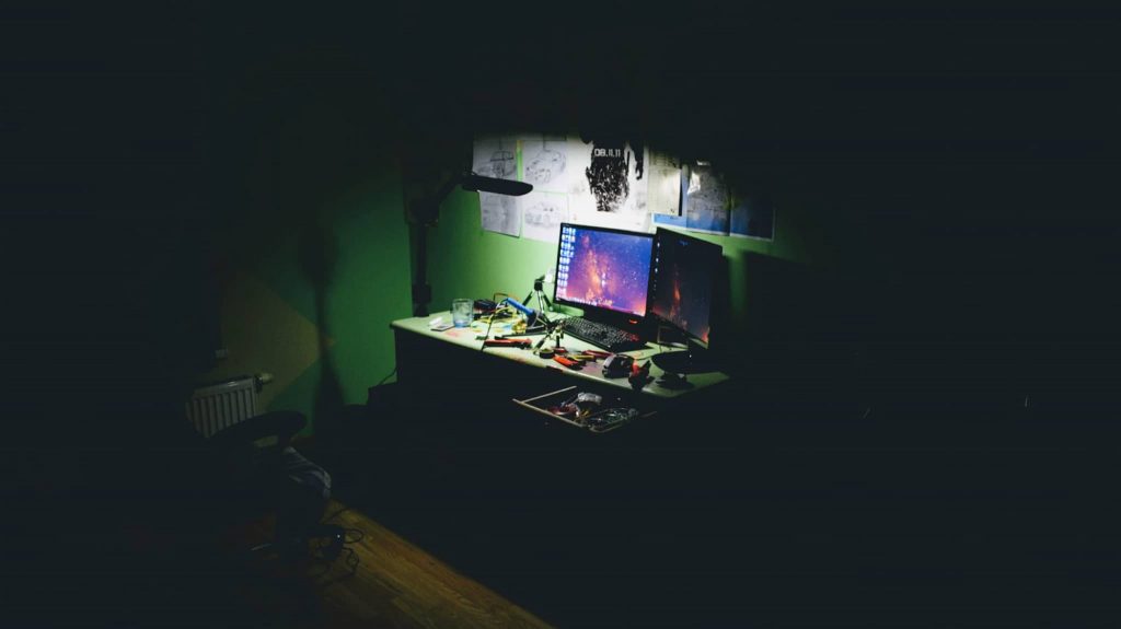 an ominous dark room with a single light shining on a desk with a computer mounted on it 